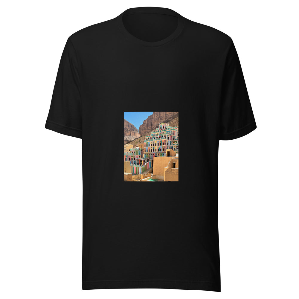Maglietta unisex nera vista di fronte. Facciate verdi, rosa e crema si alzano tra pareti rocciose color ocra, sopra tetti e scale in primo piano. Mockup del capo completo.