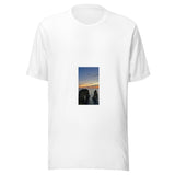 Maglietta unisex bianca vista di fronte. Due scogli emergono dal mare: quello a sinistra è attraversato da un arco, mentre sopra l’orizzonte arancio vola la piccola sagoma di un aereo. Mockup del capo completo.